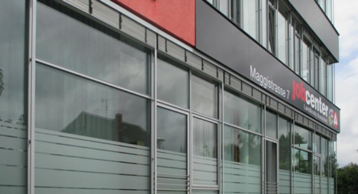 geschaeftsstelle-singen Außenansicht der Jobcenter-Geschäftsstelle Singen in der Maggistraße 7 mit großen Fensterflächen und grau-roter Fassade.